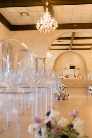 Elegant wedding venue interior with crystal chandelier, clear ghost chairs lining the aisle, and purple floral arrangements.