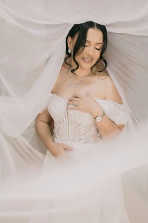 Bride in off-shoulder lace wedding gown wrapped in flowing white veil, smiling softly with elegant jewelry.