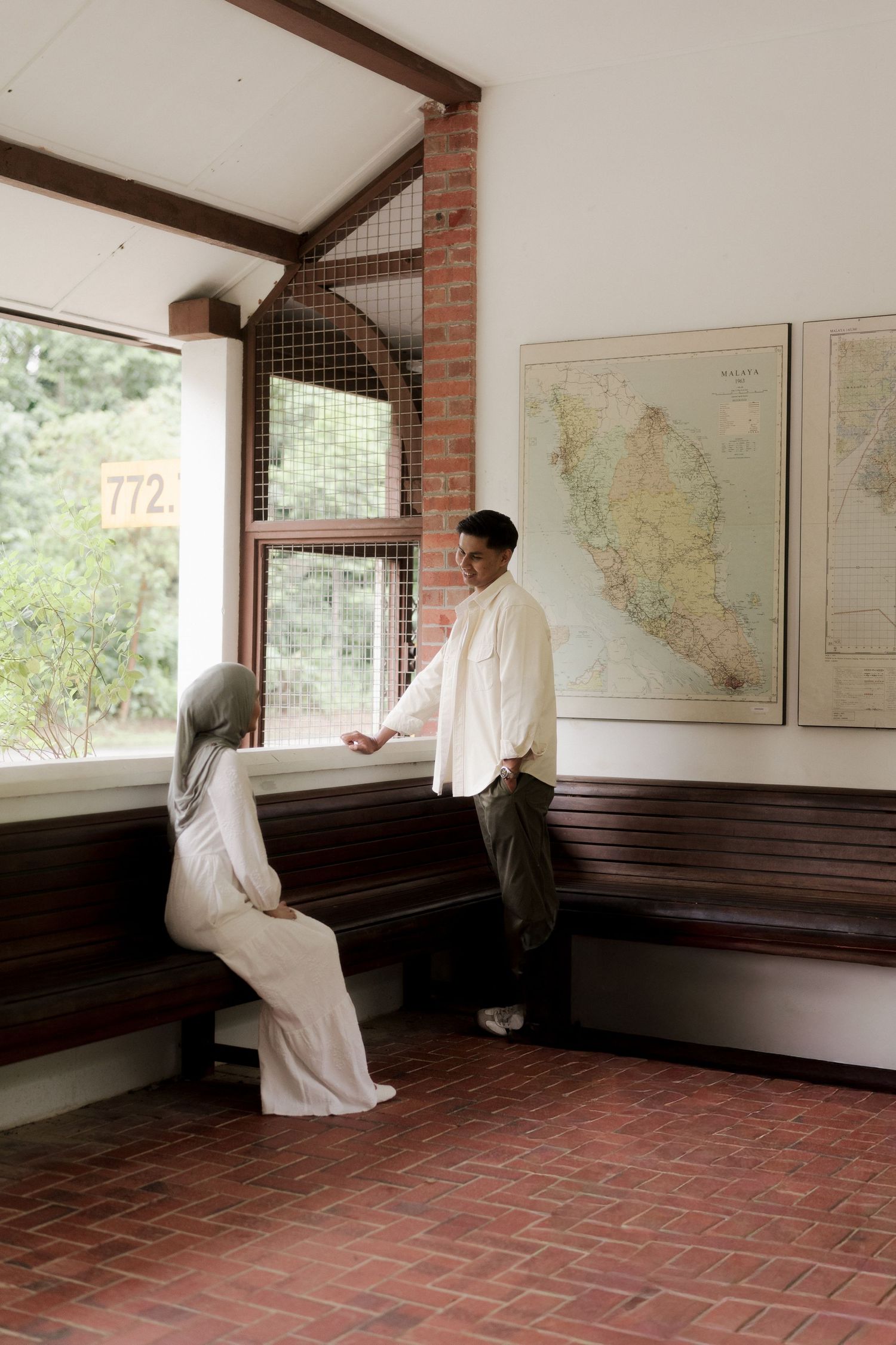 A couple in a vintage waiting room, woman in hijab seated on wooden bench, man in white shirt leaning against wall near a map.