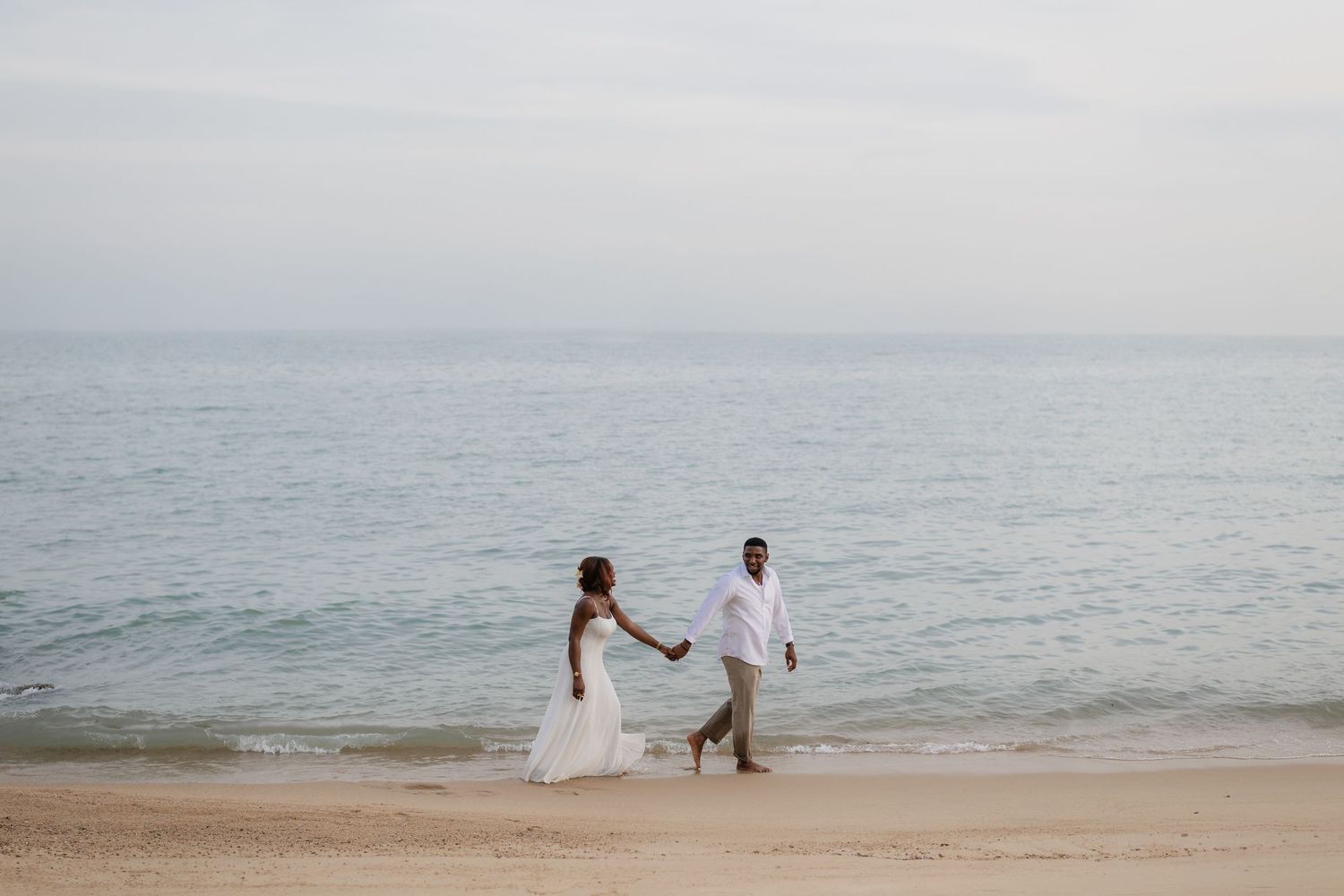 Bride and groom holding hands walking along a sandy beach with calm ocean waves in the background.