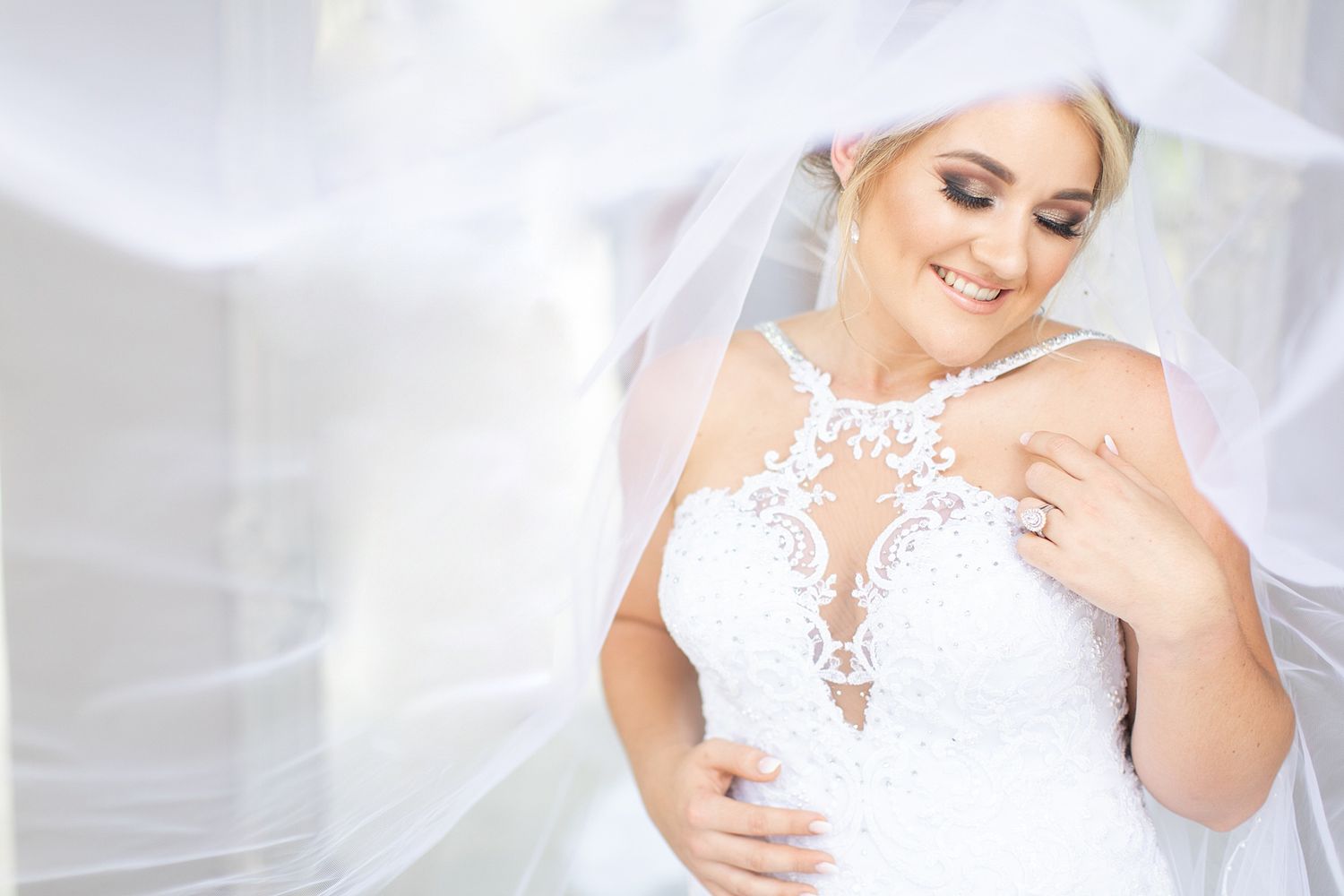 Elegant bride in off-shoulder lace wedding gown wrapped in flowing veil in soft natural light.