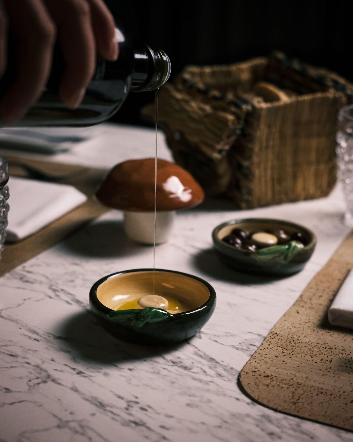 Dark ceramic bowl with egg yolk and herbs being drizzled with oil on marble surface beside mushroom decor.