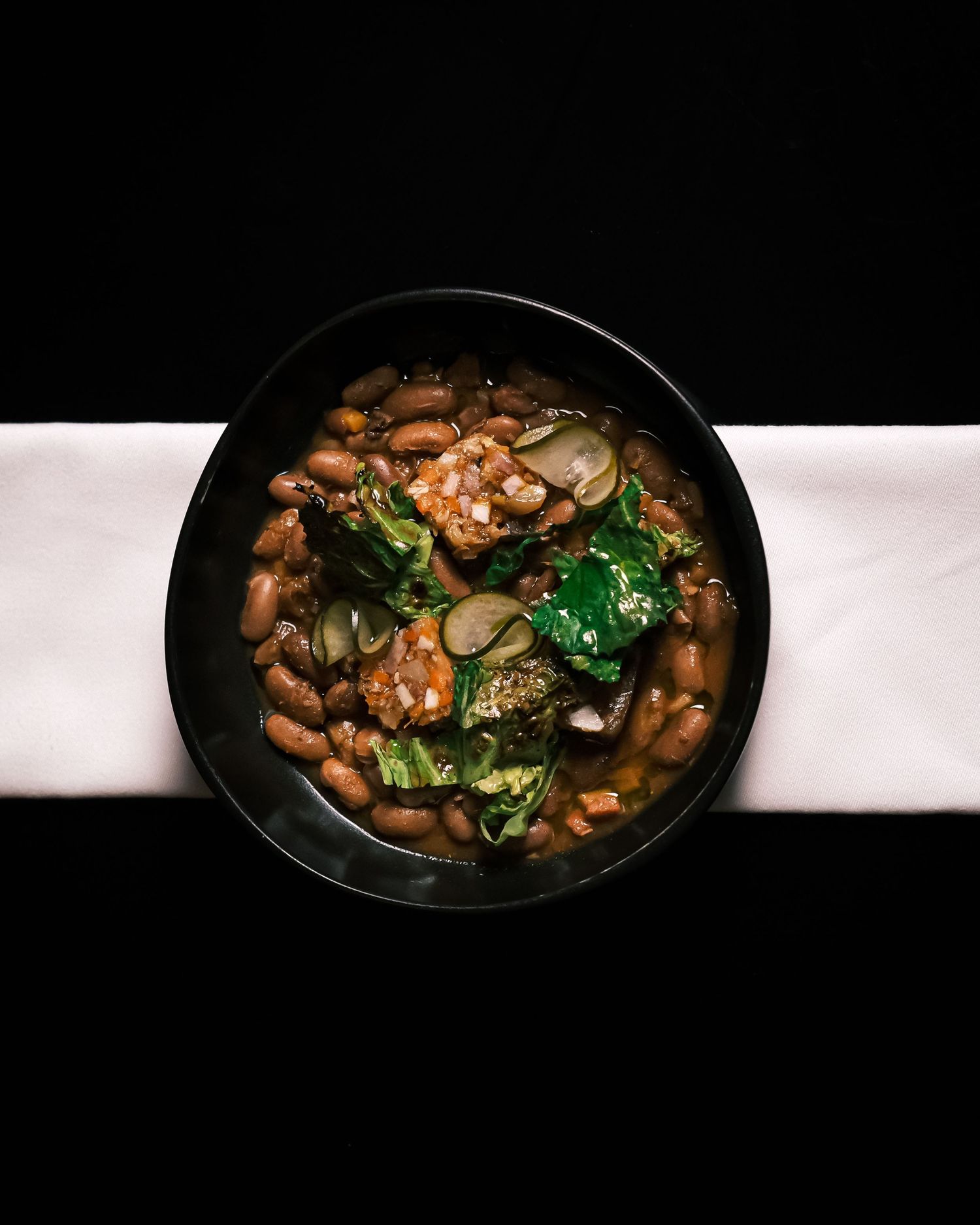 A black bowl filled with hearty bean stew with greens and vegetables, served on a white napkin.