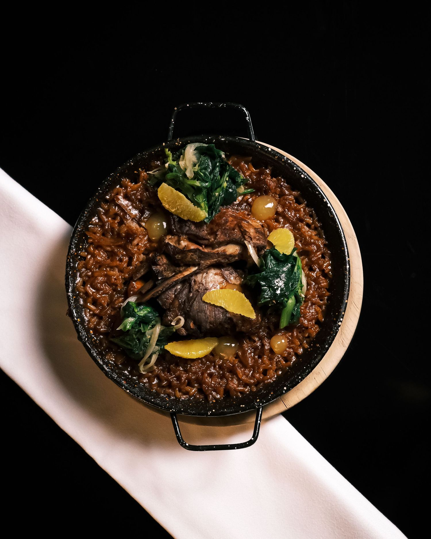 A paella pan filled with rich brown rice, braised meat, sautéed greens, grapes, and citrus slices on a white cloth.