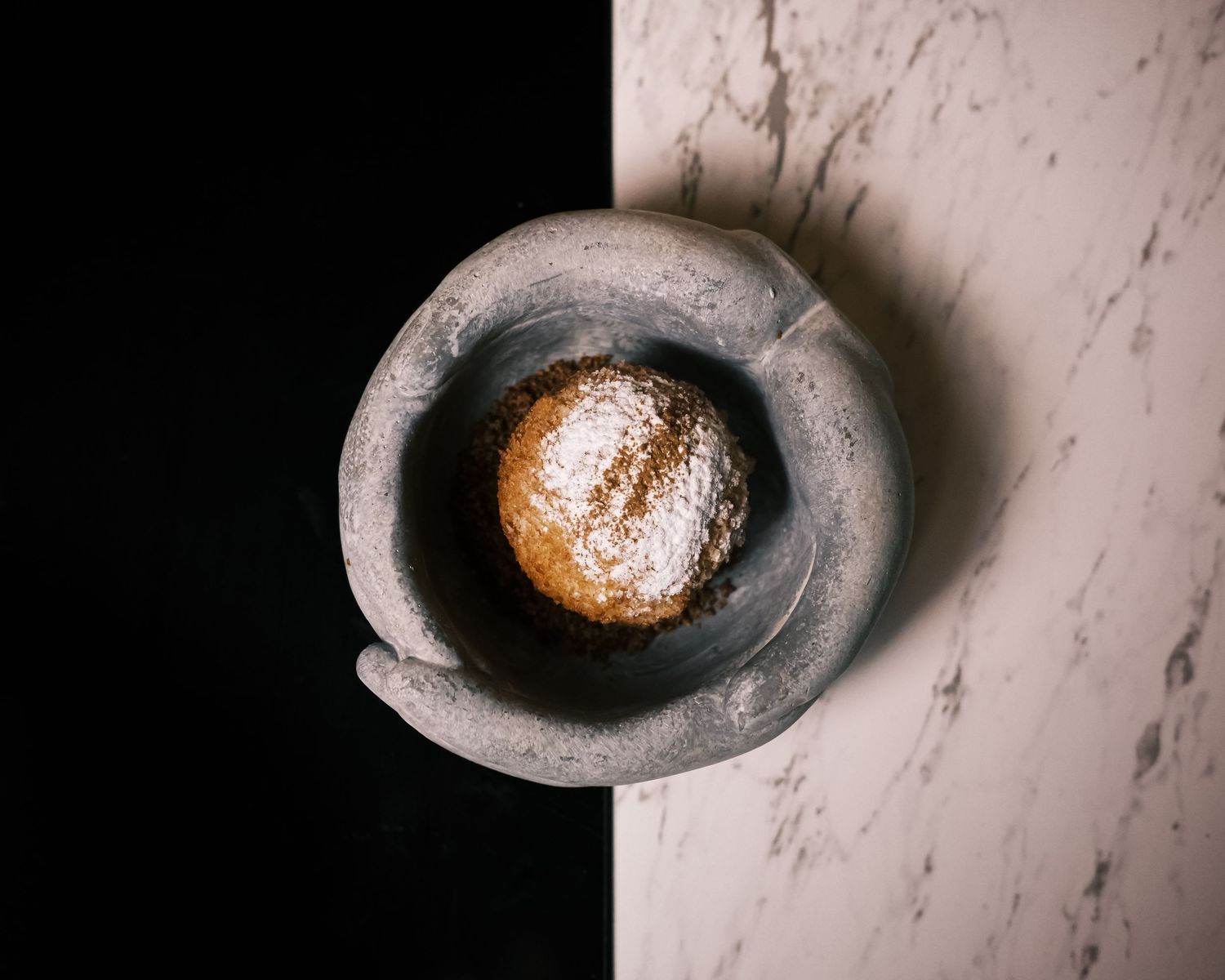 A powdered sugar-dusted round pastry resting in a stone mortar on a marble surface.