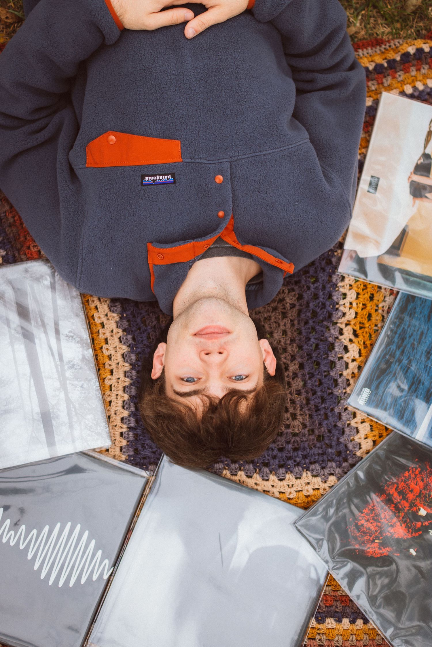 A young man lies on a retro crocheted blanket and smiles softly up at the camera with his vinyl record collection spread out around him.