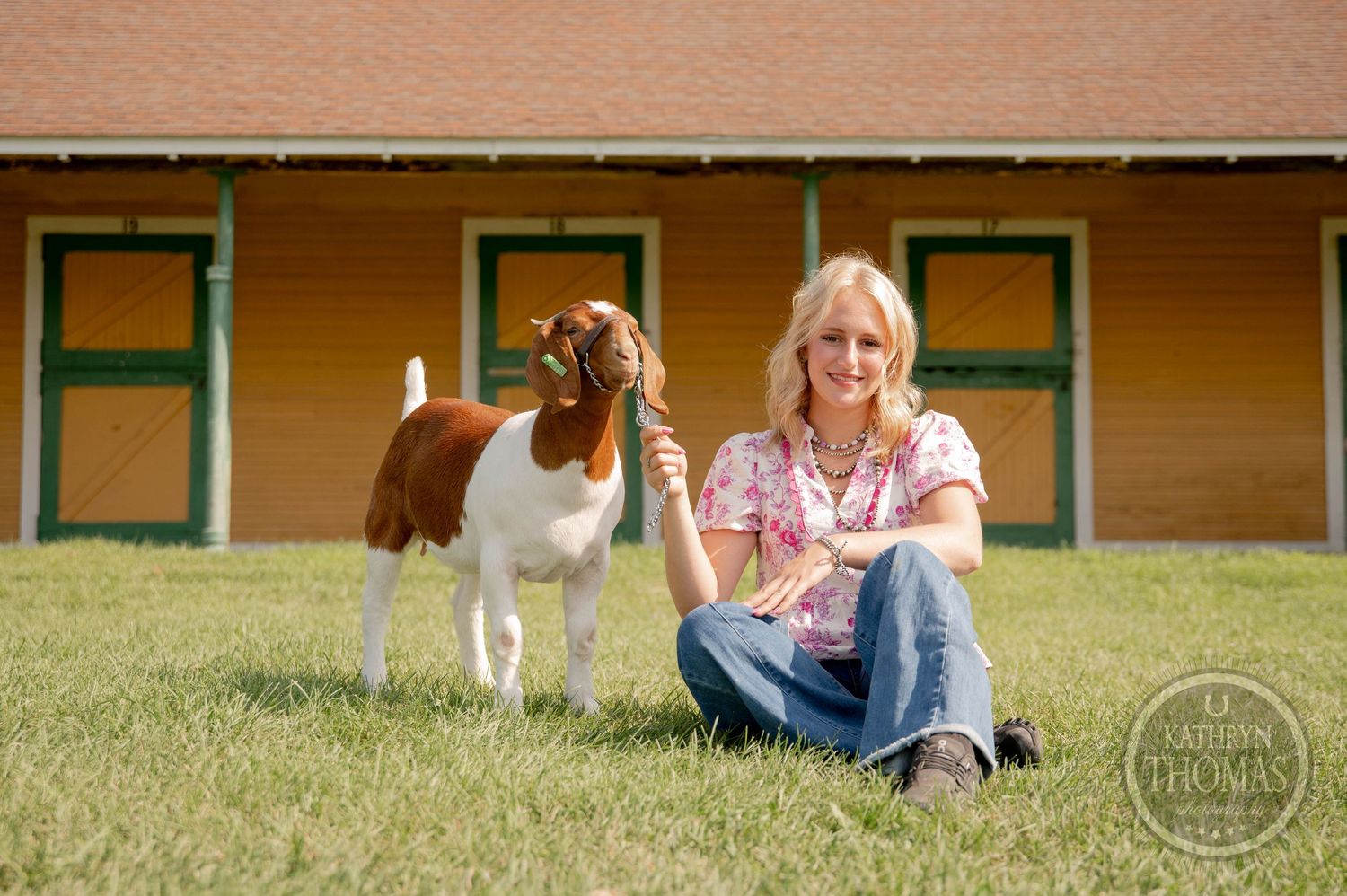 Smiling blonde woman sitting on grass with a brown and white goat in front of a yellow barn.
