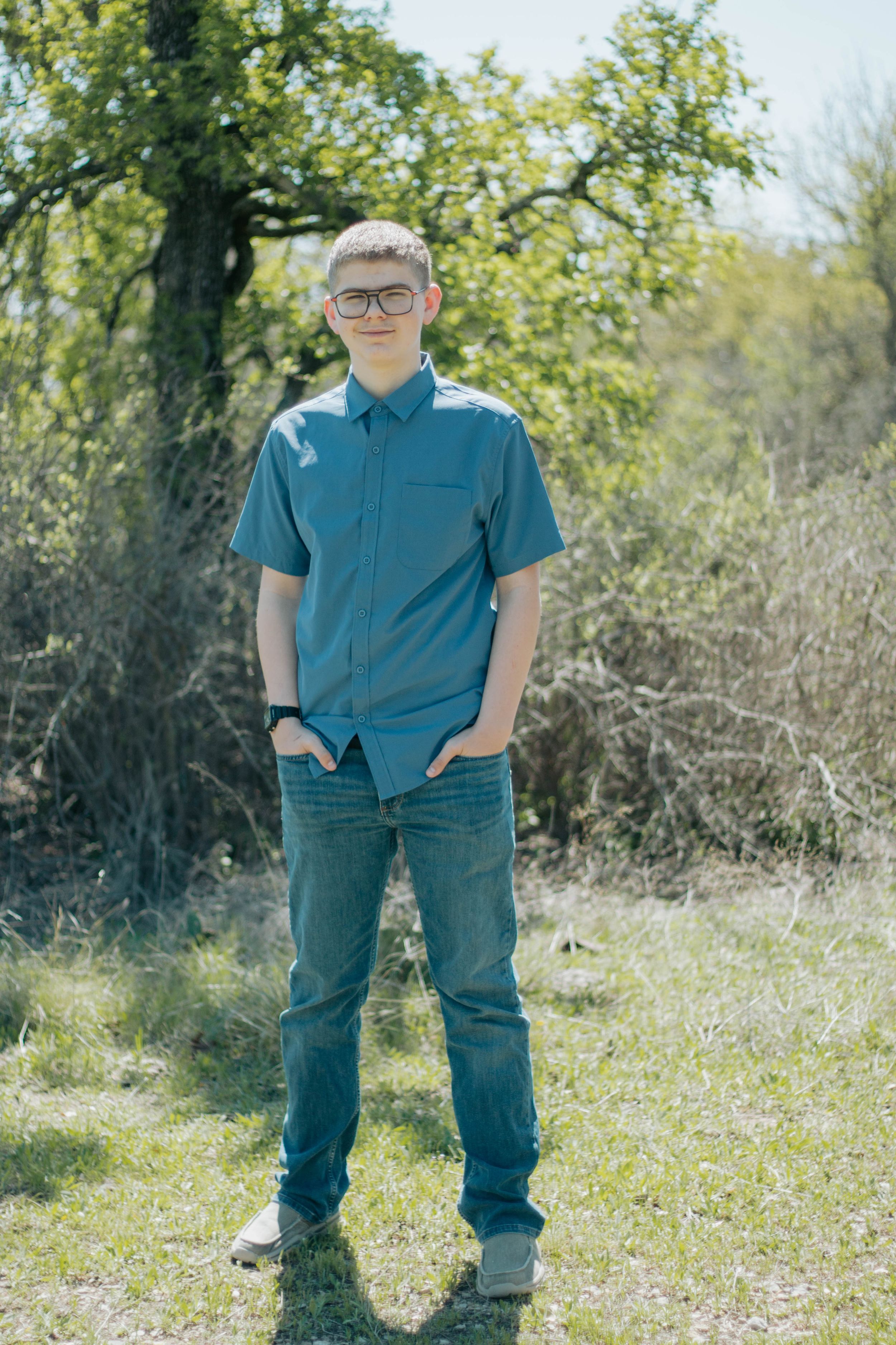 Teen boy in blue shirt and jeans with glasses stands outdoors on grass with trees in background.