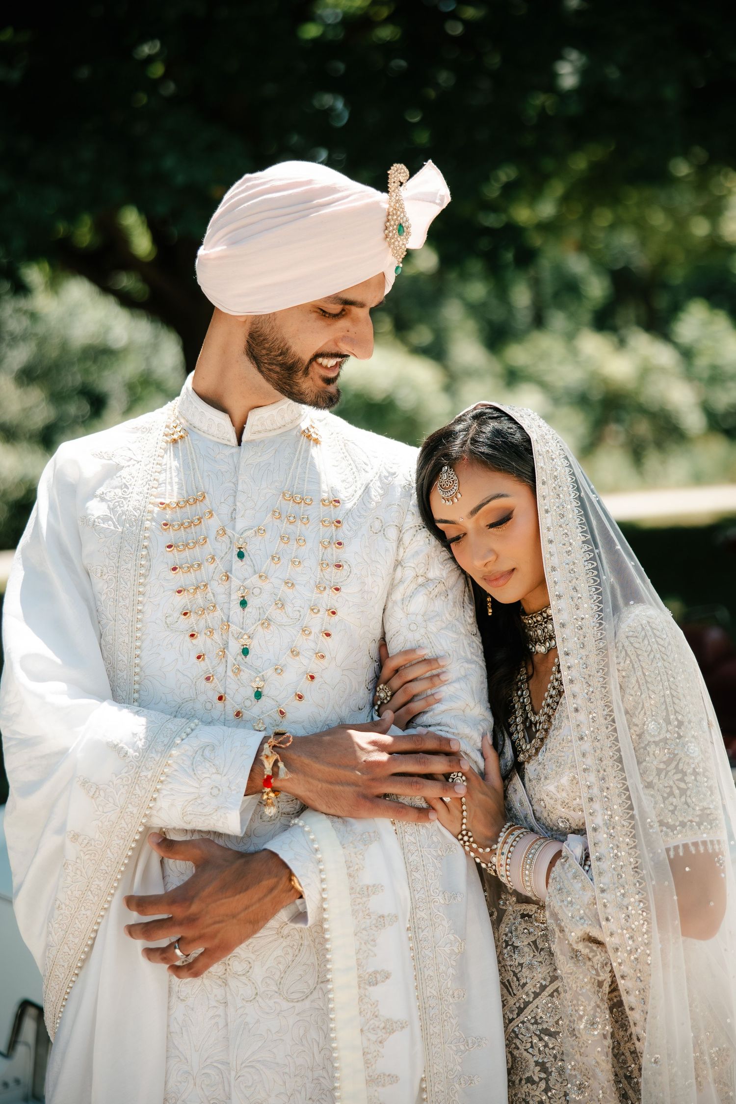 A Sikh couple in traditional white wedding attire embraces outdoors, the groom in a turban and sherwani, bride in an embroidered lehenga.