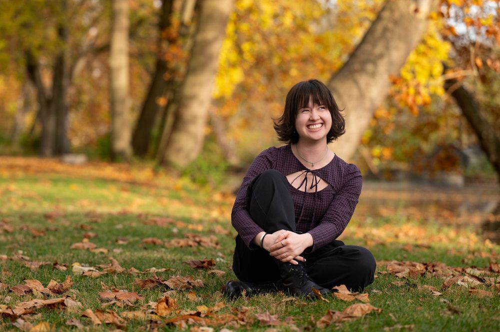 Outdoor autumn portrait of high school senior in a purple top sitting on fallen leaves with golden trees in background.