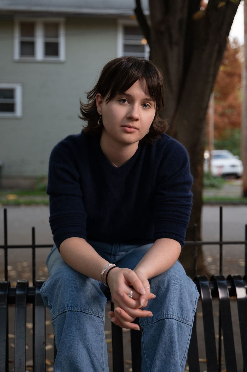 Casual portrait of high school senior in navy sweater and jeans sitting on metal railing with house in background.