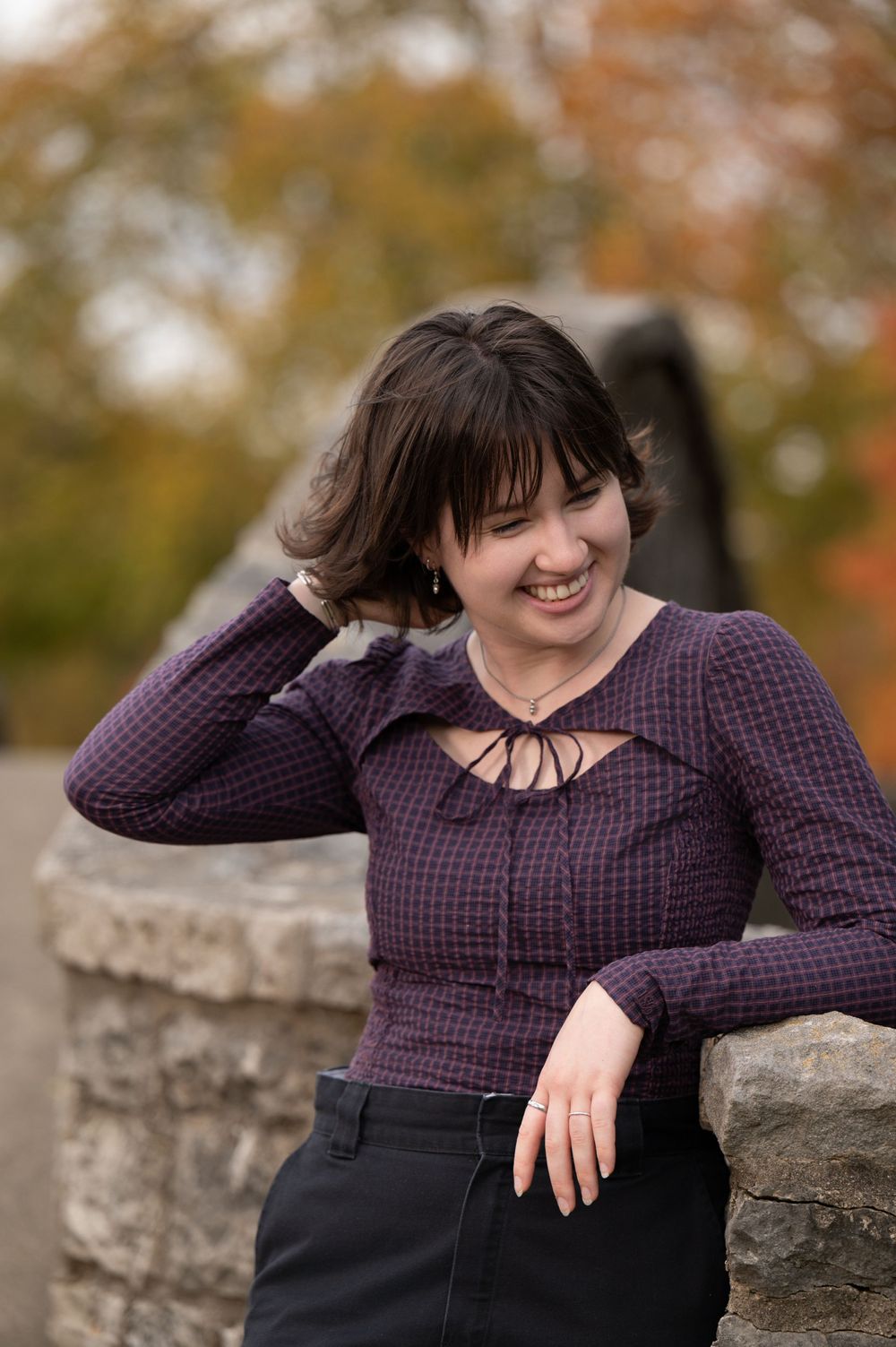 Portrait of high school senior wearing a purple knit top leaning on a stone wall with autumn foliage behind.