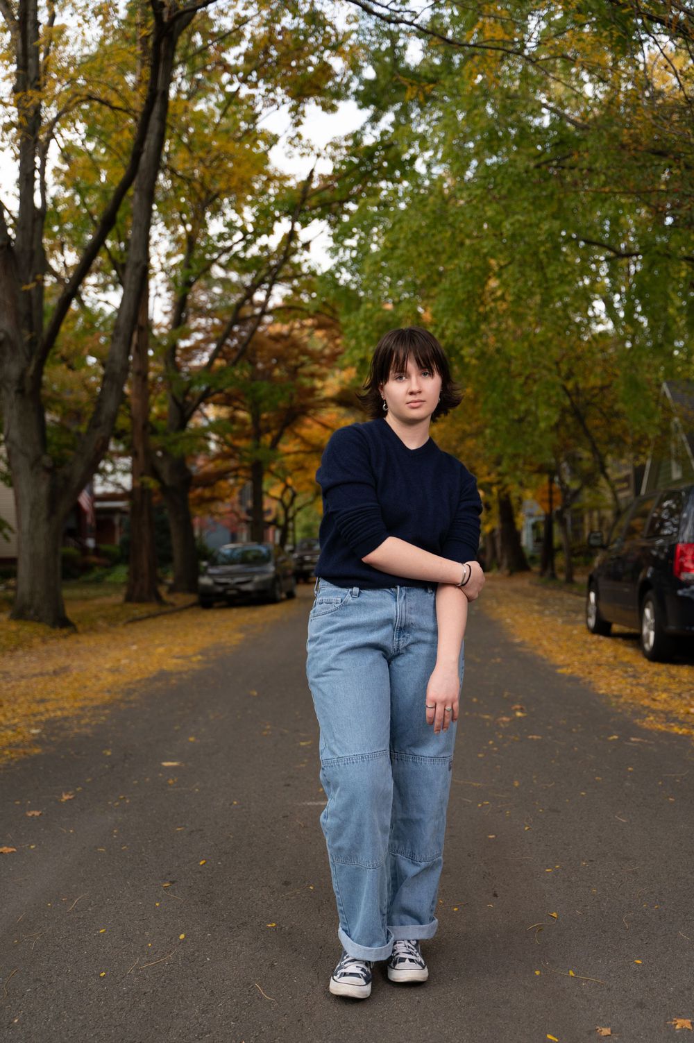 Full-length photo of high school senior in jeans and navy sweater standing on tree-lined street during fall.