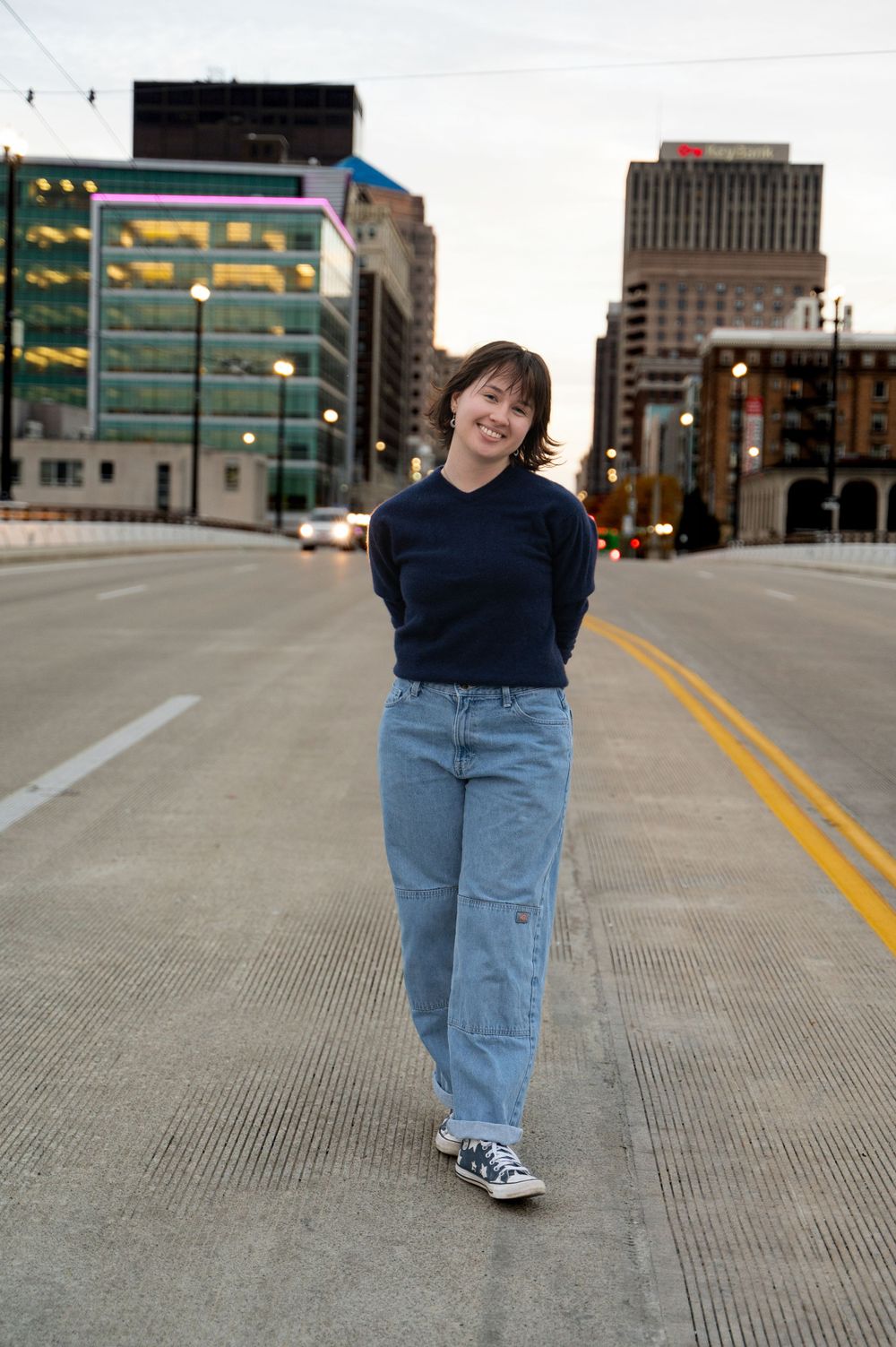 Urban street photo of high school senior in blue jeans and navy sweater with city buildings in background.