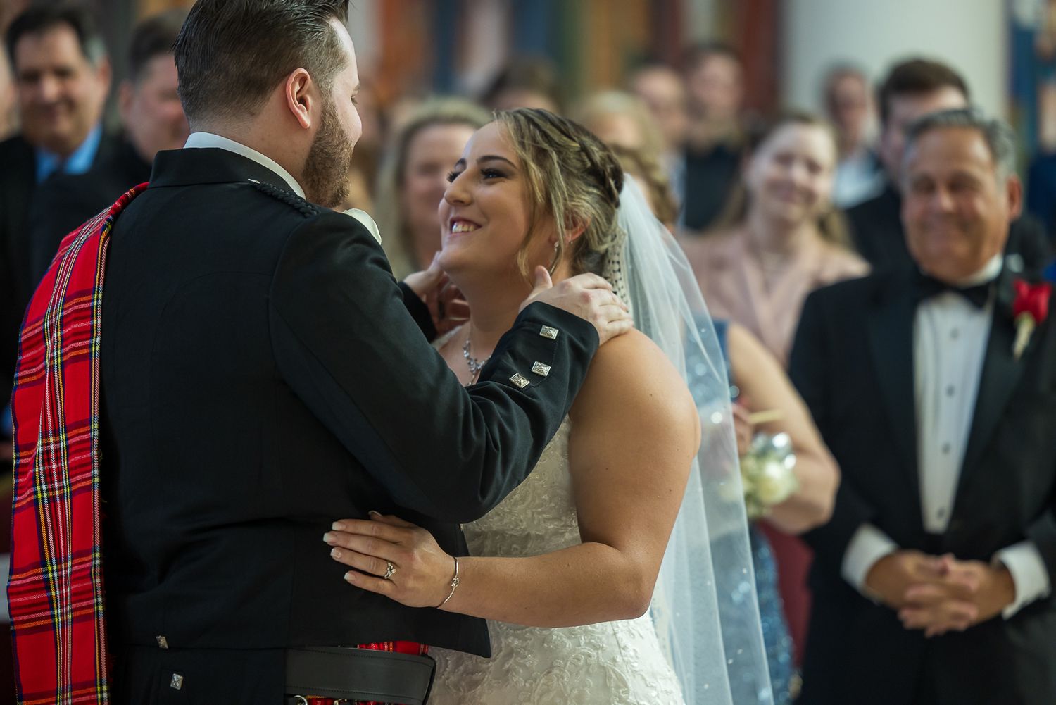 Bride and groom share a joyful moment at the altar, groom in Scottish tartan kilt, bride in lace wedding gown with veil.