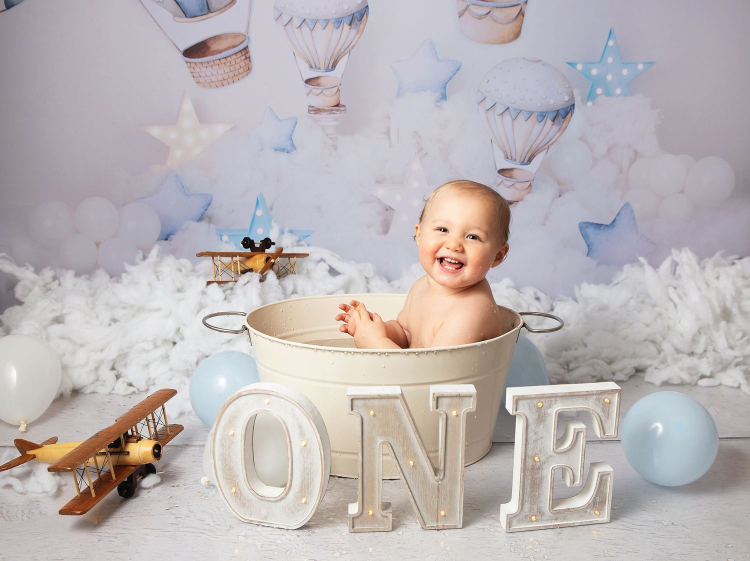 Smiling baby sitting in a tin bucket surrounded by ONE letters, toy airplane, and balloons for a first birthday cake smash photo.