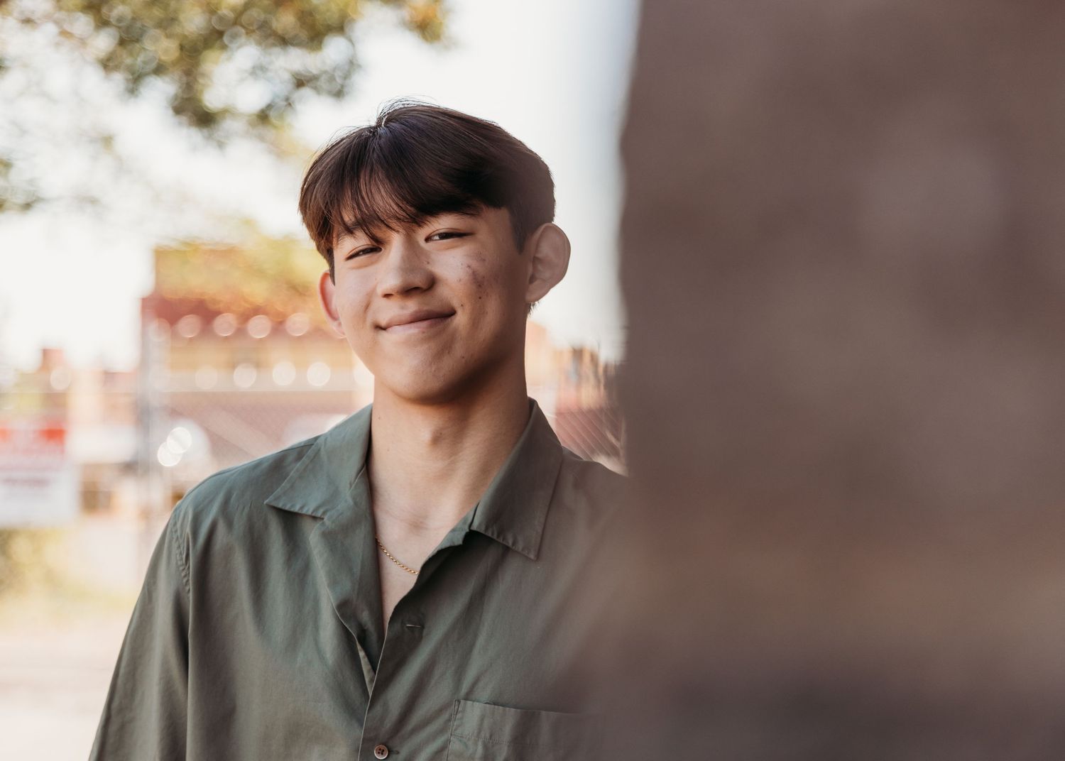 Smiling young Asian man in olive green shirt and cap posing outdoors in warm sunlight.