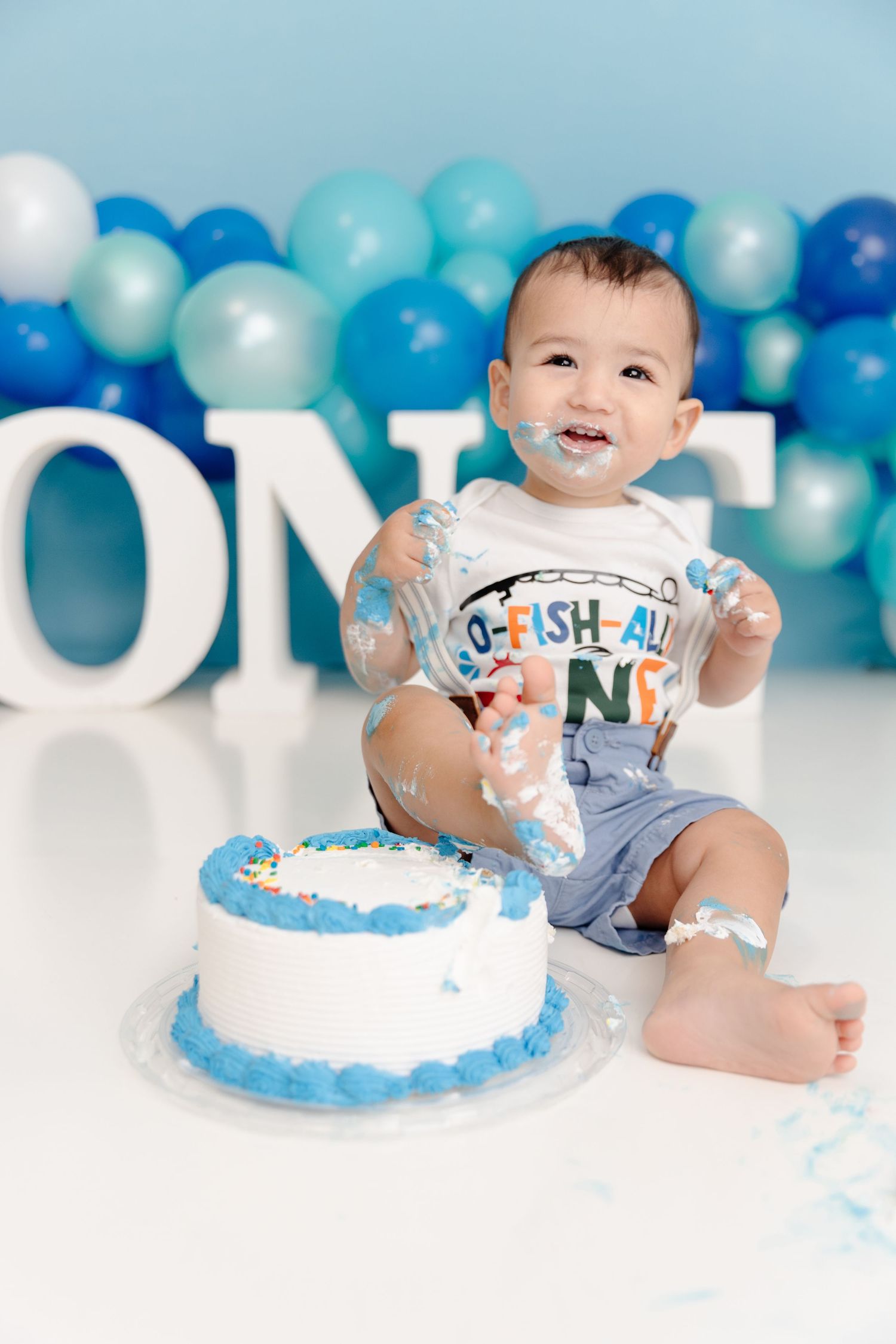 Baby boy smashing first birthday cake surrounded by blue balloons and ONE letters in a festive photo session.