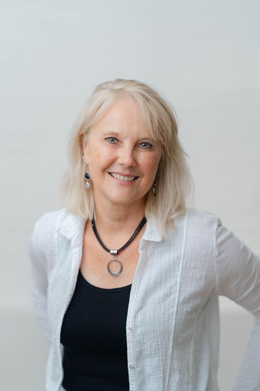 Smiling middle-aged blonde woman wearing a white linen shirt and black necklace against a light background.
