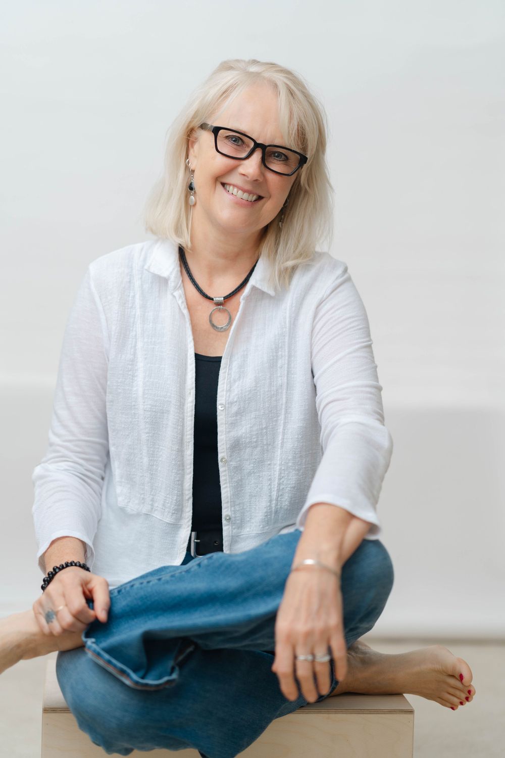 Cheerful blonde woman with black glasses sitting cross-legged, wearing white shirt and jeans, smiling warmly.