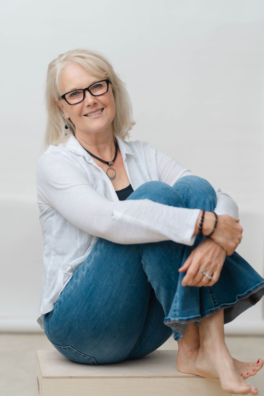 Relaxed blonde woman in glasses and white shirt sitting barefoot on a wooden block, hugging her knees.