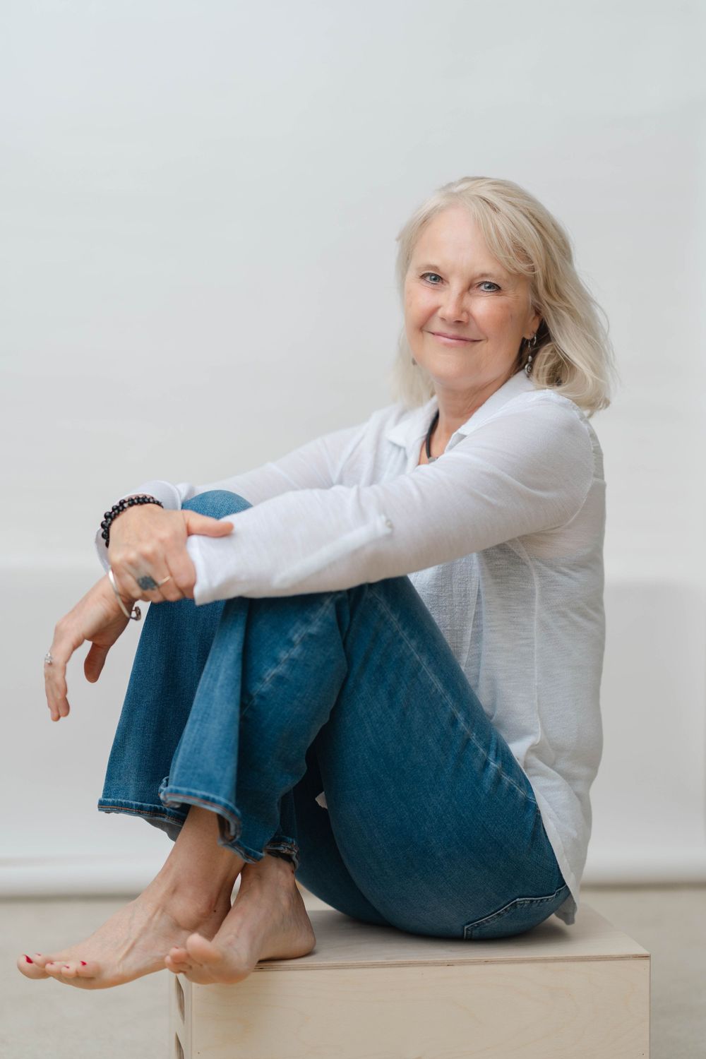 Smiling middle-aged woman with blonde hair sitting barefoot on a box, wearing white shirt and jeans.