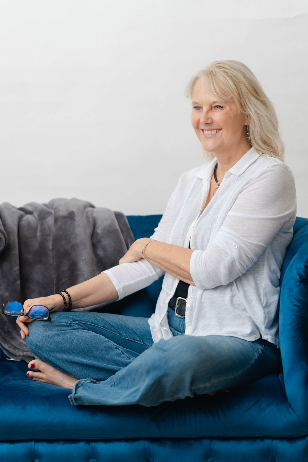 Happy blonde woman sitting cross-legged on a blue velvet sofa, holding glasses, wearing white shirt and jeans.