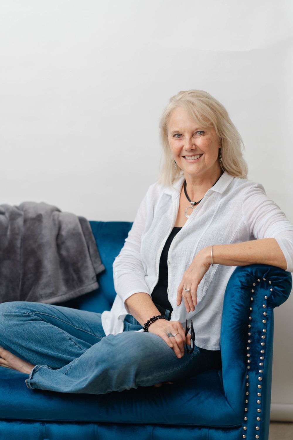Smiling blonde woman relaxing cross-legged on a blue velvet sofa, wearing a white shirt and jeans.
