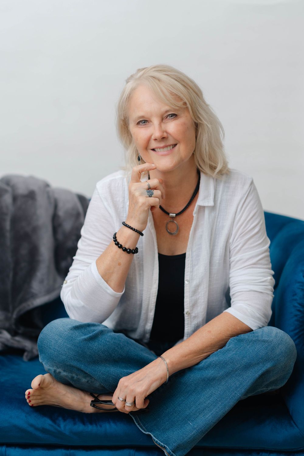 Blonde woman sitting cross-legged on a blue couch, smiling warmly in a white shirt and jeans.