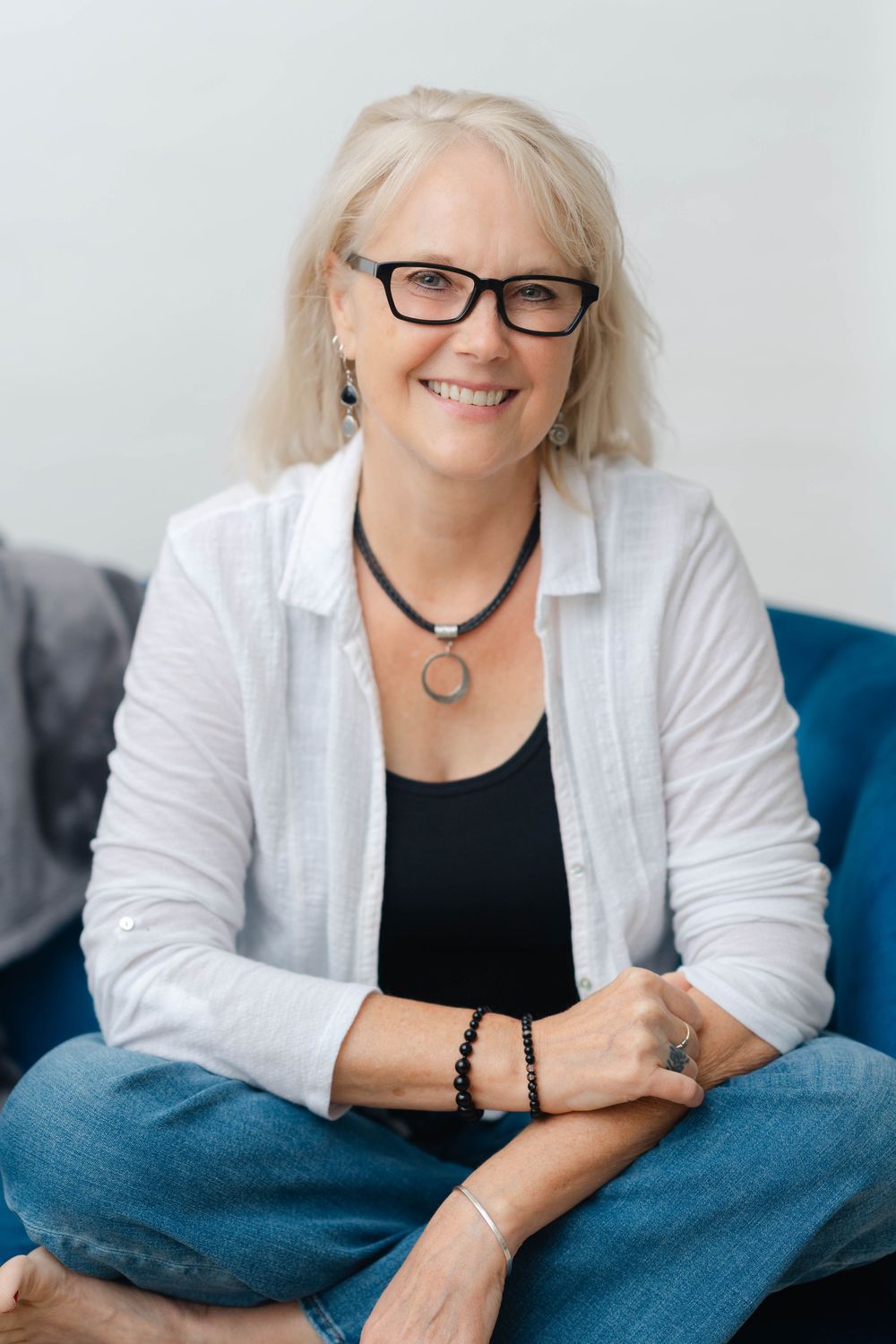 Smiling middle-aged woman with gray hair and glasses, wearing white shirt and jeans, sitting cross-legged on blue sofa.