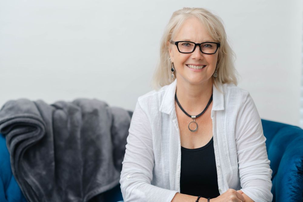 Smiling middle-aged woman with gray hair and glasses wearing a white cardigan, seated on a blue couch.