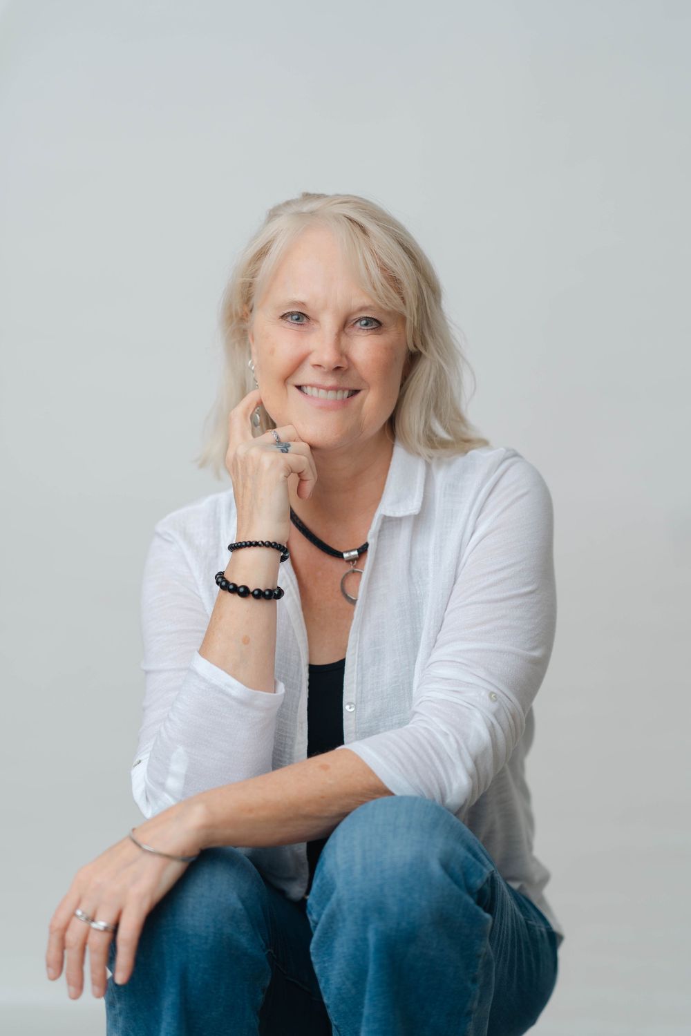 Smiling blonde woman in white cardigan and jeans sitting casually with hand on chin, wearing black bracelet.