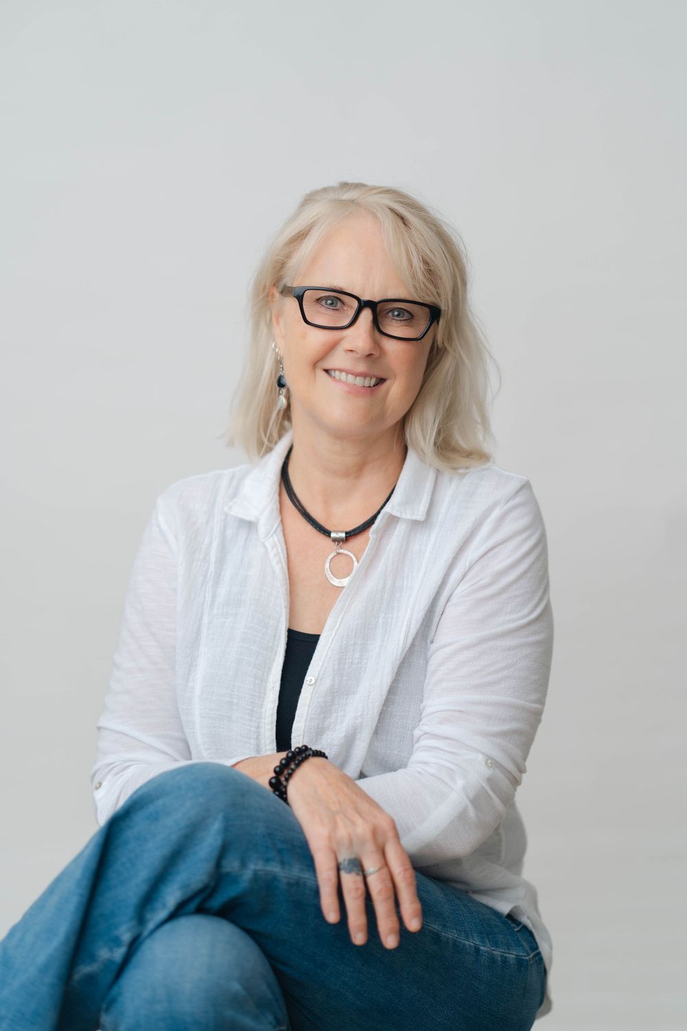 Smiling middle-aged woman with blonde hair and glasses wearing a white linen shirt and jeans, sitting casually.