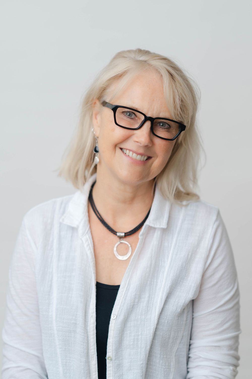 Smiling middle-aged woman with blonde hair, black glasses, and a white linen shirt wearing a circular pendant necklace.