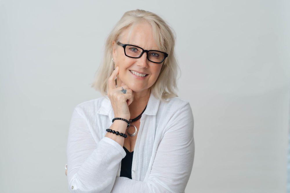 Smiling senior woman with gray hair and glasses posing confidently in a white blazer against a light background.