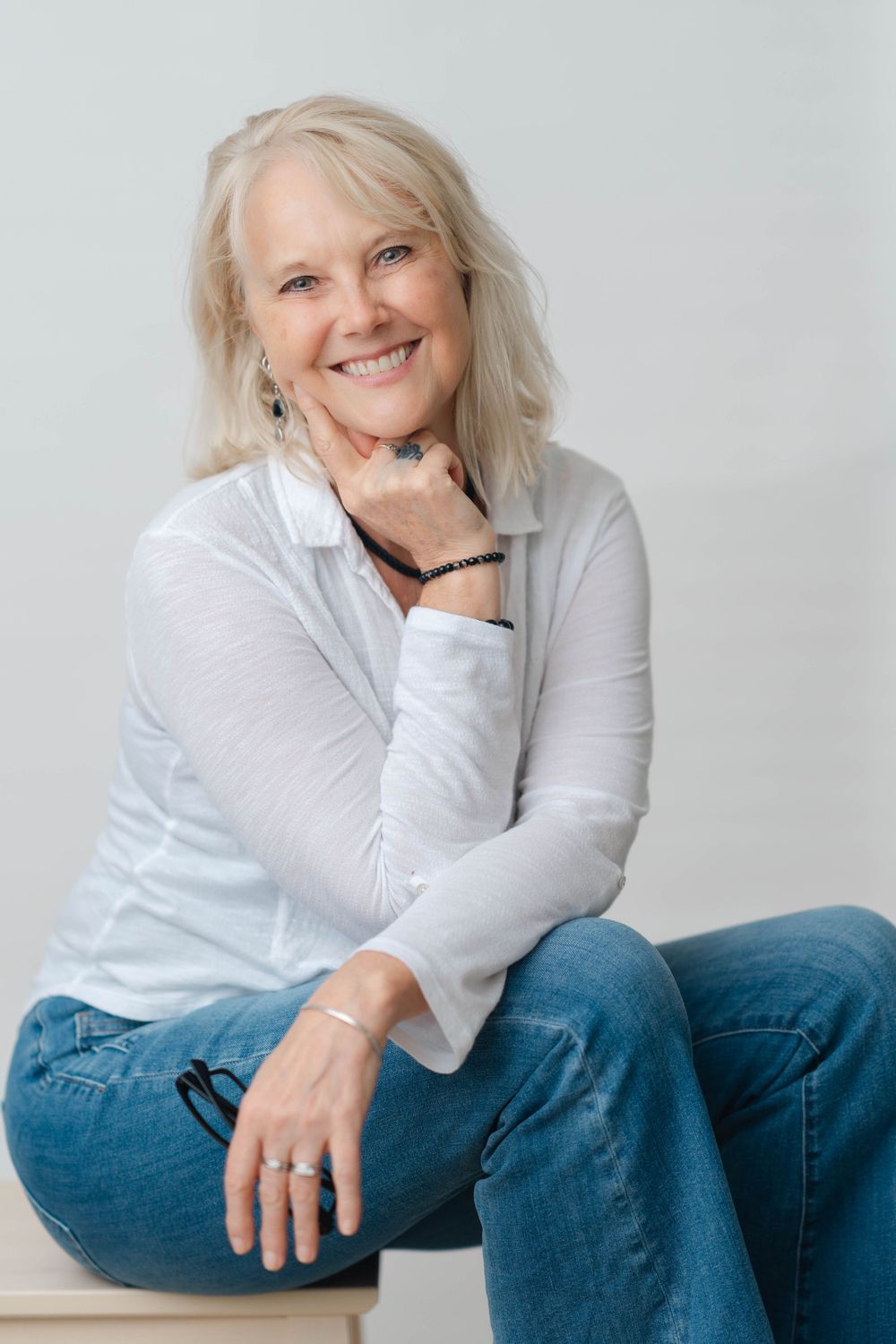 Cheerful older woman with blonde hair in white top and jeans, sitting relaxed with hand on chin, smiling warmly.