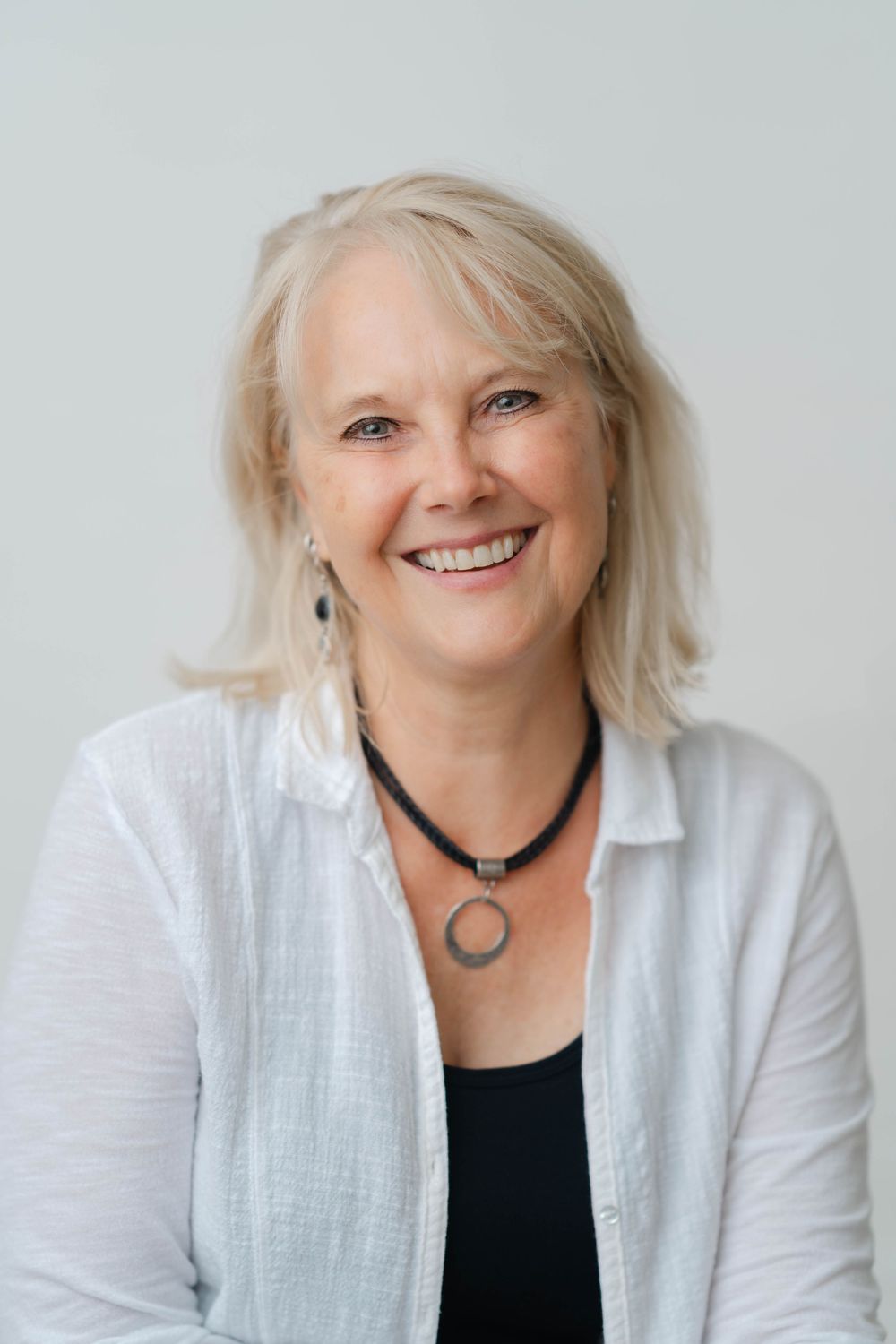 Happy middle-aged blonde woman smiling warmly, wearing a white linen shirt and black necklace against a light background.