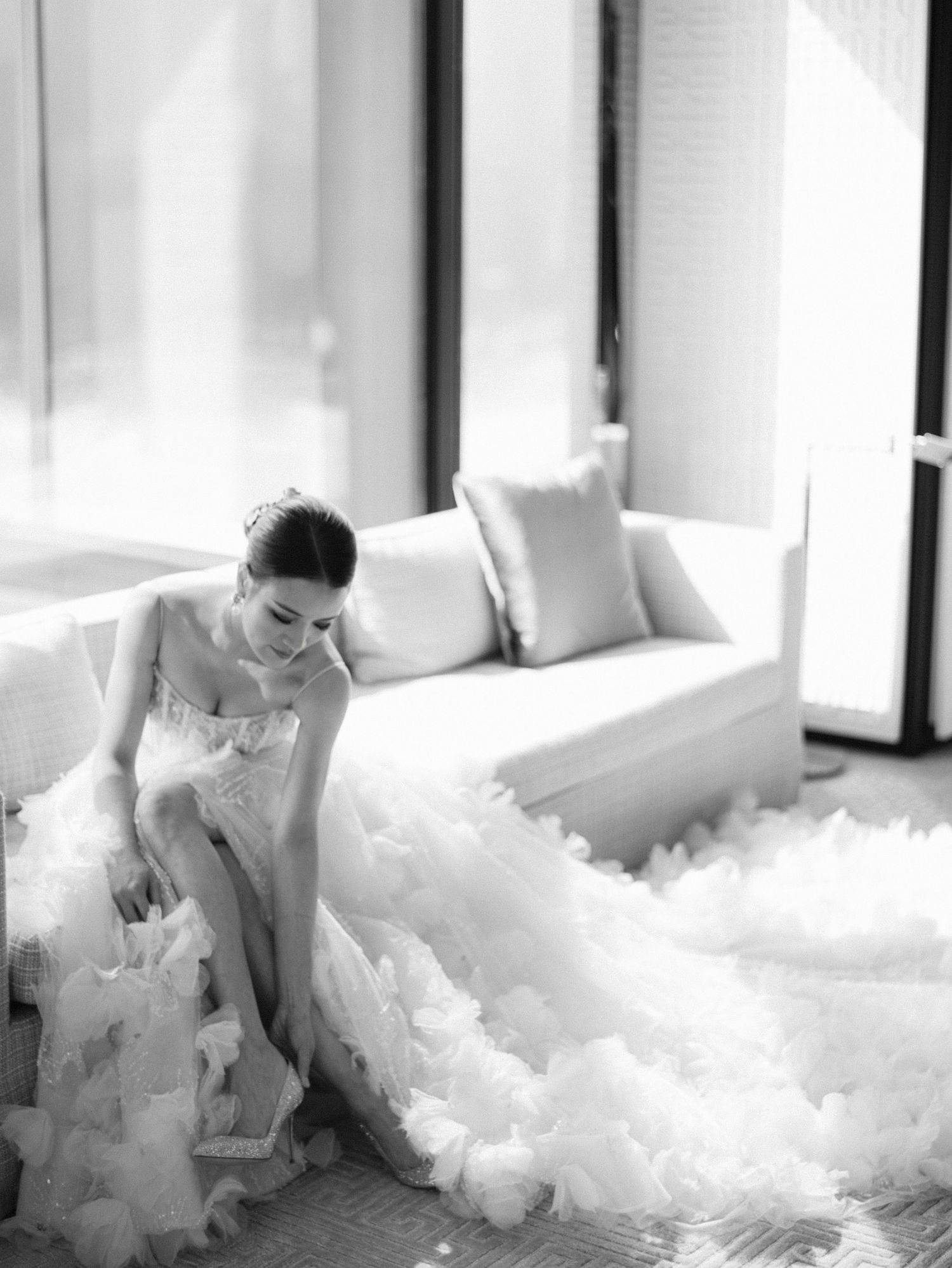 Bride in a voluminous ruffled white wedding gown seated on floor in black and white photo.