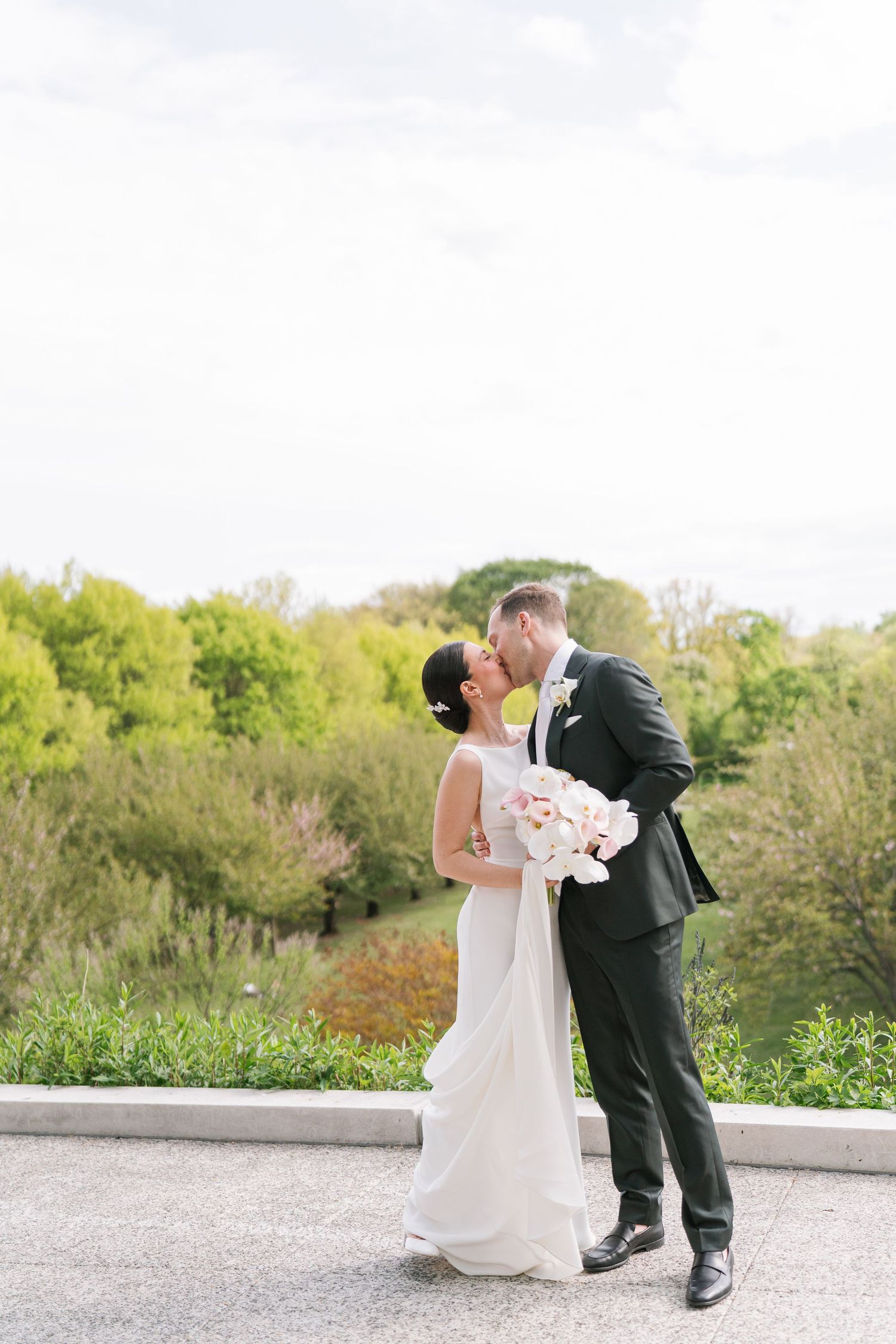 A bride and groom share a romantic kiss outdoors, surrounded by lush green trees, as she holds a white floral bouquet.