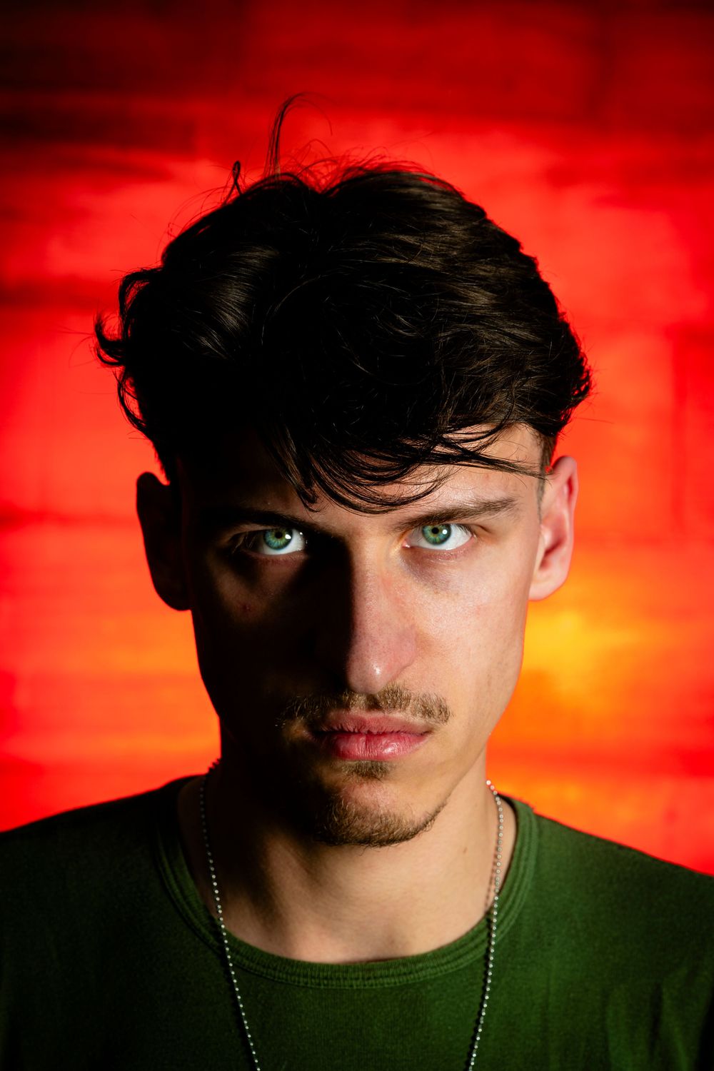 Young man with blue eyes and dark curly hair poses against a vibrant red-orange brick wall background.