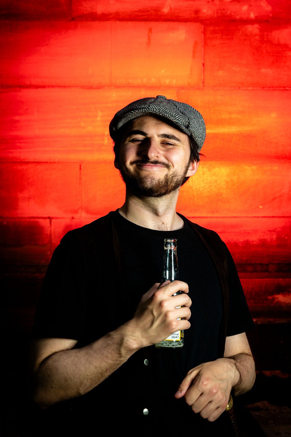 Smiling bearded man in flat cap holding beer bottle against vibrant red wall.