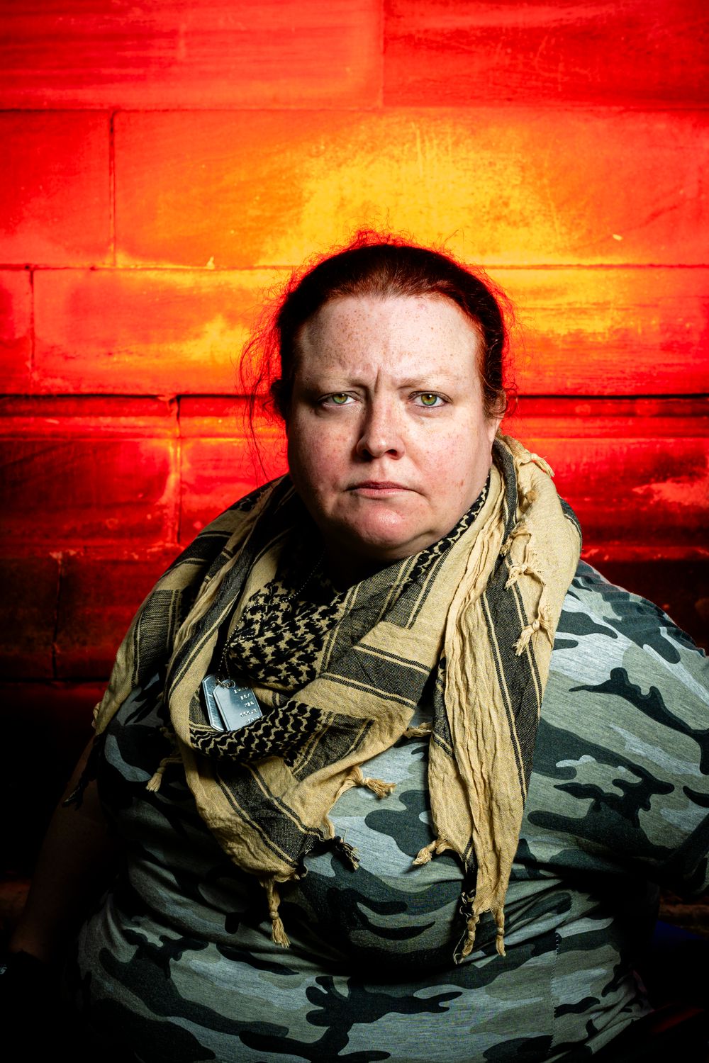 Woman in camouflage jacket and scarf against red brick wall.