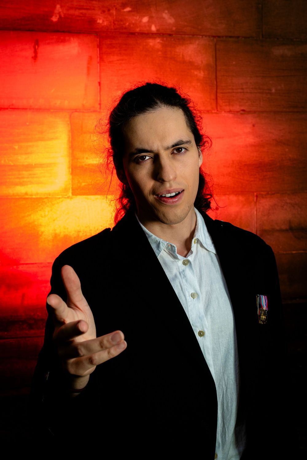 Man with long dark hair in black blazer gesturing against red brick wall.