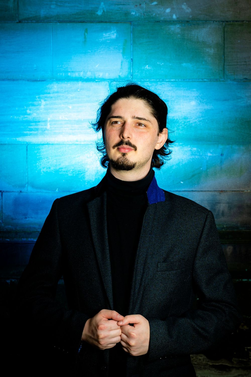 Young man in black turtleneck and blazer against blue wall.