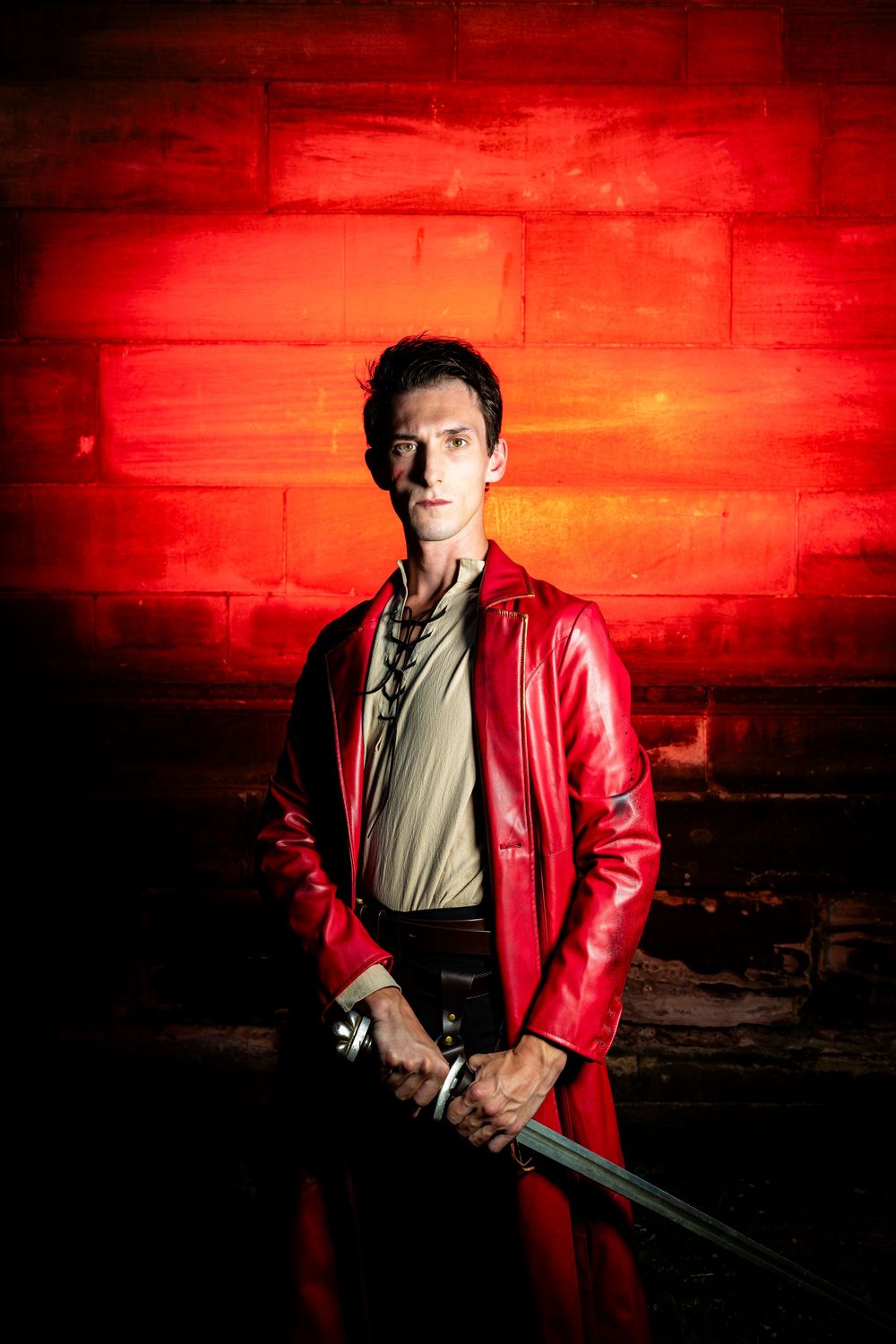 Man in red leather coat holding sword against red brick wall.