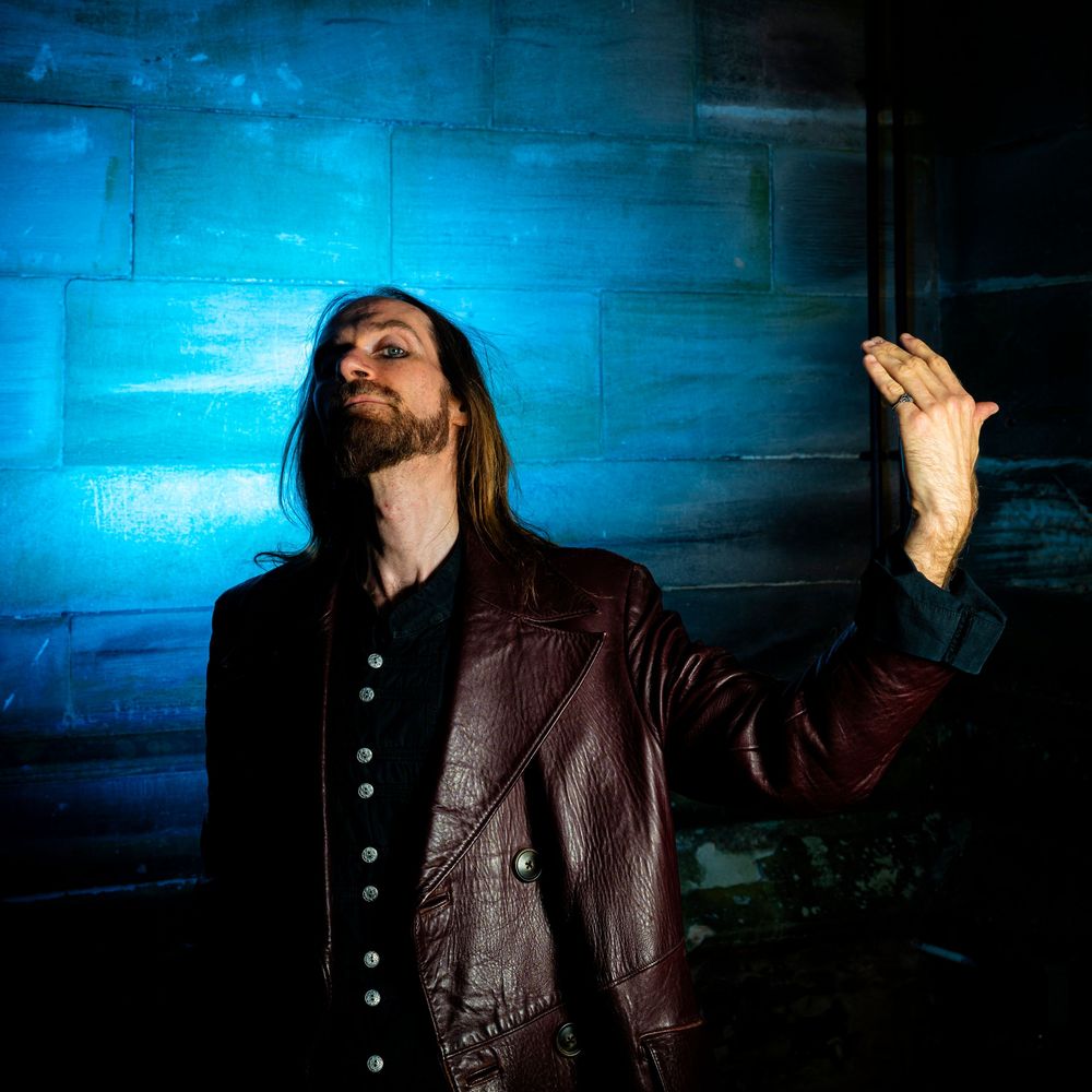 Long-haired man in burgundy leather jacket posing dramatically against blue wall.