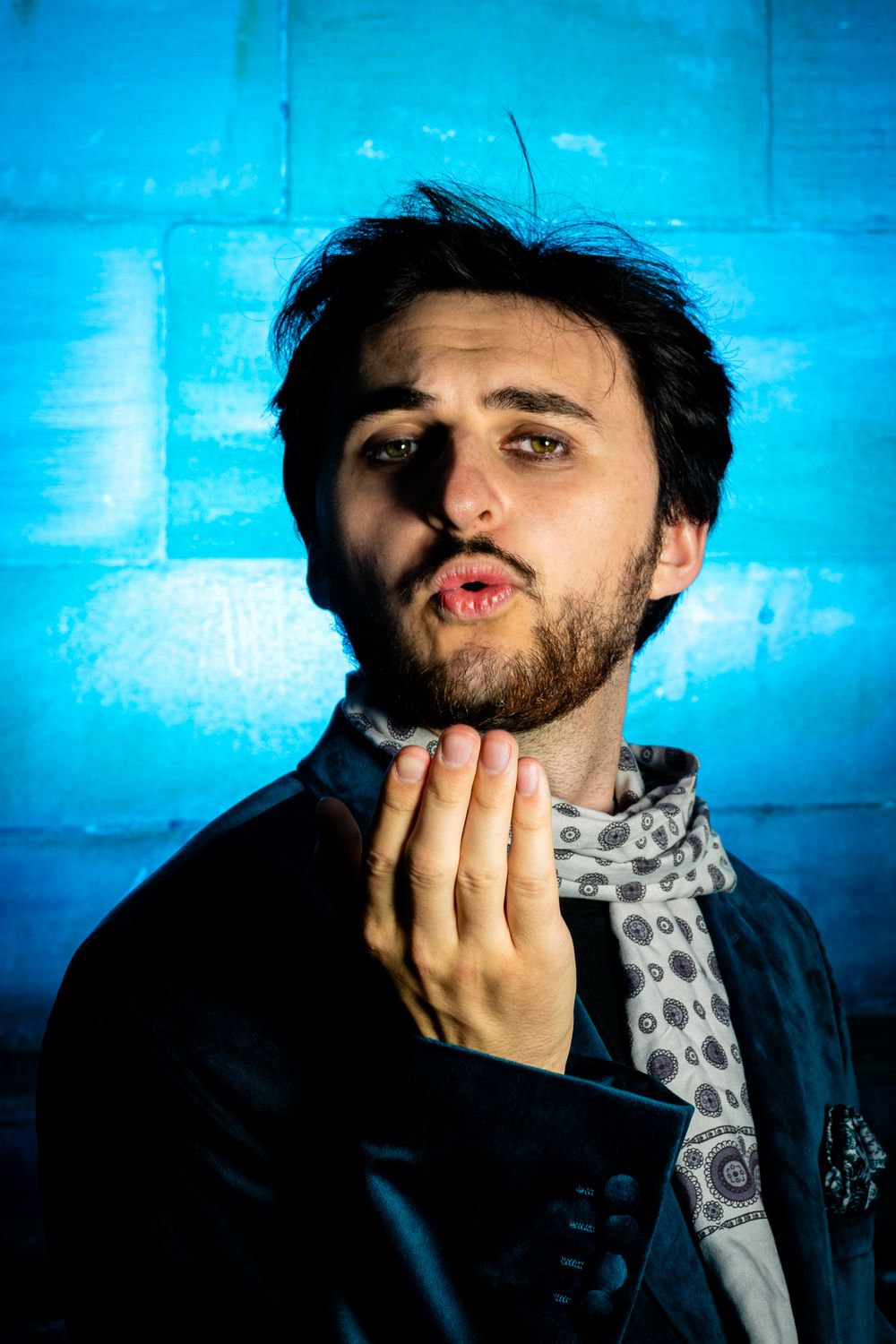 Bearded man in black jacket with scarf against blue brick wall.