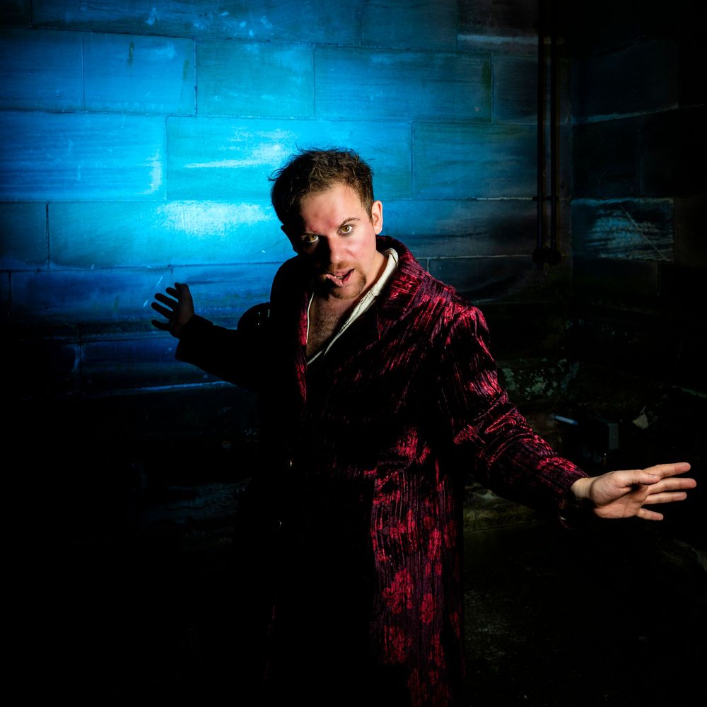 A dramatic male performer in a red velvet robe poses against a blue-lit brick wall in a theatrical setting.