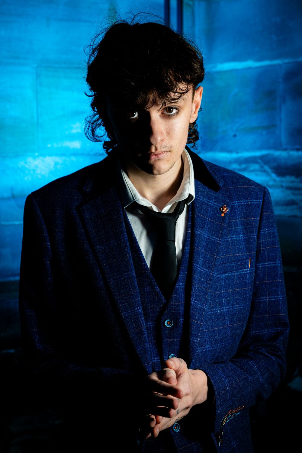 Young man in blue plaid suit with tie posing in dramatic blue lighting.