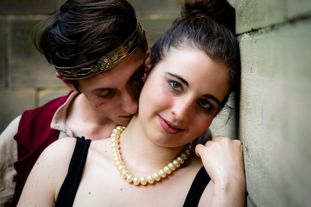 Young couple posing together, woman wearing pearl necklace against stone wall.